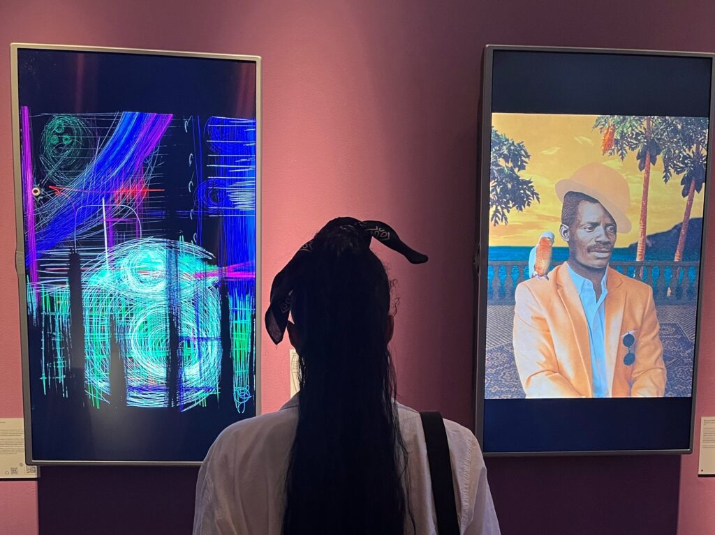 A visitor seen from behind stands in front of two large framed artworks on a deep pink gallery wall. The left work is a dark, neon-lit abstract digital piece with layered geometric patterns in blue, green, and purple. The right is a vivid figurative painting of a man in an orange blazer with a parrot on his shoulder, set against a warm tropical sunset backdrop.
Photo credit: Carla Candace via Dupe Photos