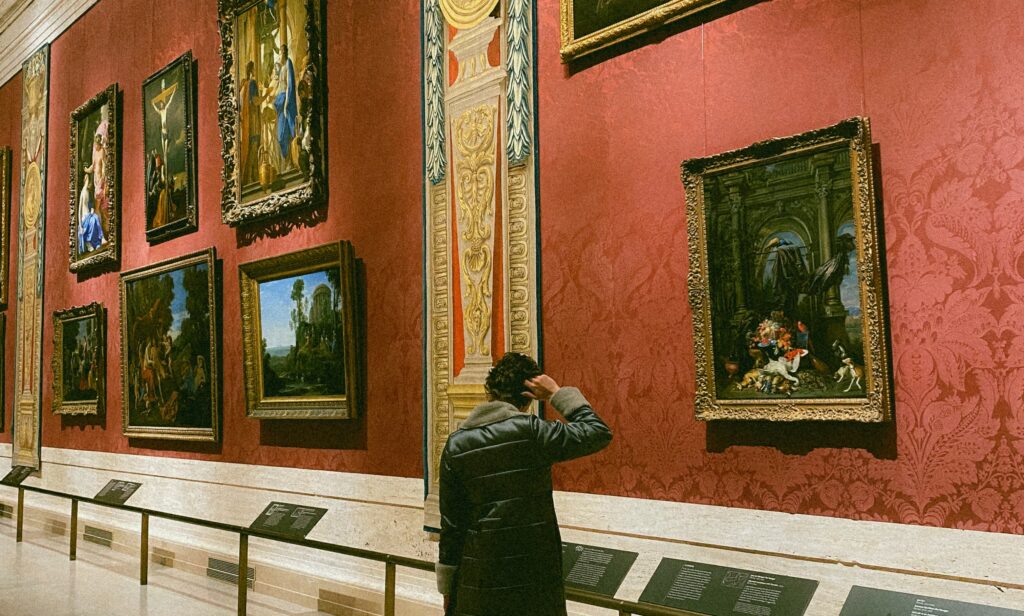 A visitor stands alone in a grand museum gallery, studying a large ornate painting up close. Deep red damask walls display a salon-style arrangement of gilt-framed Old Master paintings, with interpretive labels mounted along the base of the wall below.
Photo credit: Rachel Elaine via Dupe Photos
