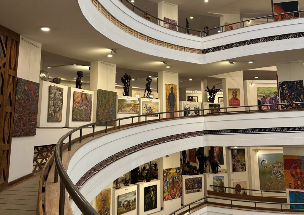 A multi-level museum gallery with a curved spiral staircase, displaying a dense collection of colorful paintings and dark bronze sculptures across two visible floors. Works feature vibrant figurative and portrait subjects with rich patterns, reflecting African artistic traditions.
Photo credit: Villa via Dupe Photos