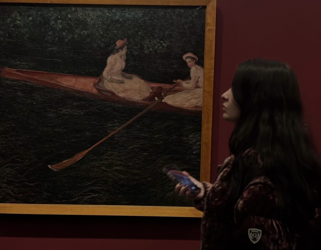 A young woman holding a phone stands in profile in front of a large Impressionist painting on a deep red gallery wall. The painting depicts two figures in white dresses and hats seated in a rowing boat on dark water.
Photo credit: Bruna Daniela via Dupe Photos