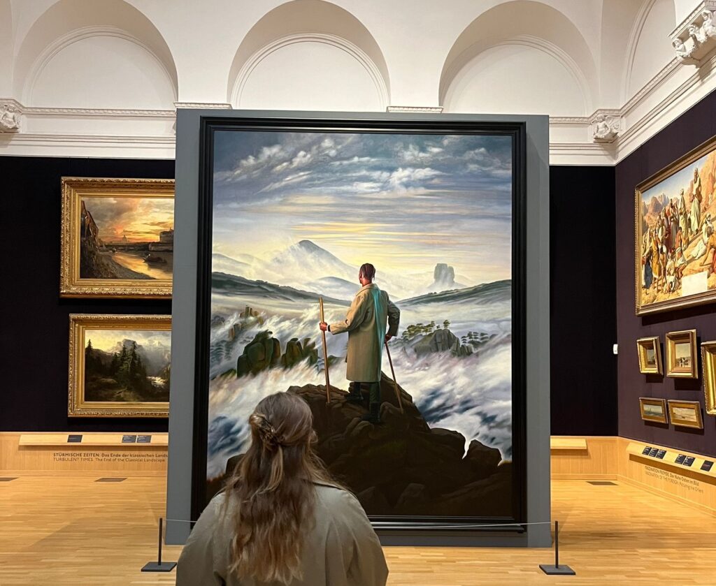 A visitor seen from behind looks up at a large-scale Romantic landscape painting mounted on a dark gallery wall. The painting depicts a lone figure standing on a rocky outcrop holding a walking staff, gazing out over a dramatic misty mountain landscape under a luminous sky. Smaller gilt-framed landscape paintings are visible on either side, with bilingual German and English section labels mounted along the gallery floor.

Photo credit: Jessie Sperling via Dupe Photos