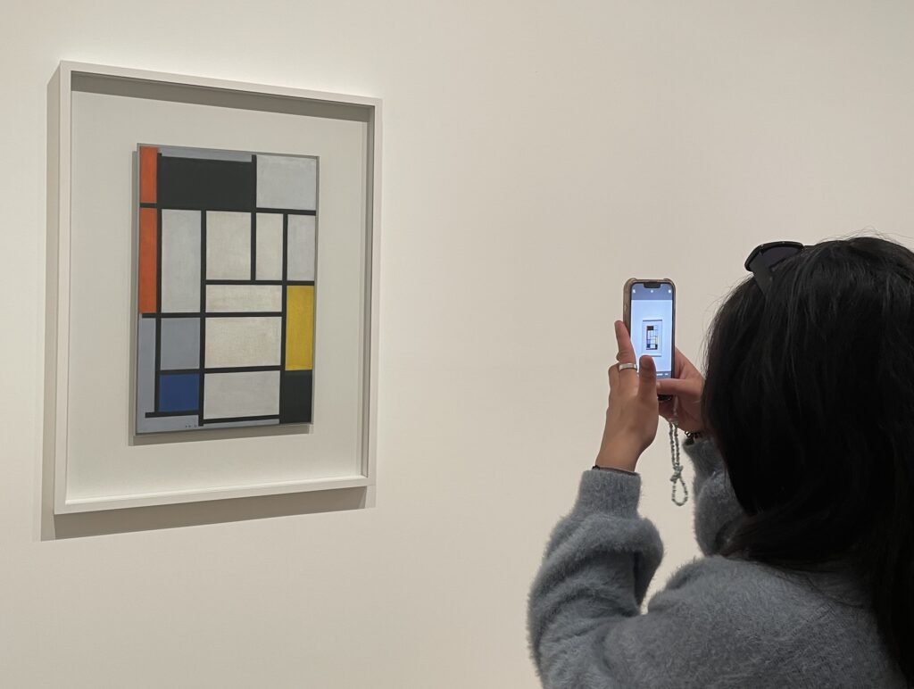 A visitor photographs a framed abstract painting on a white gallery wall using a smartphone. The painting features a grid composition of bold black lines dividing rectangular blocks of red, yellow, blue, gray, and black in the De Stijl style.
Photo credit: Reet Rachwani via Dupe Photos