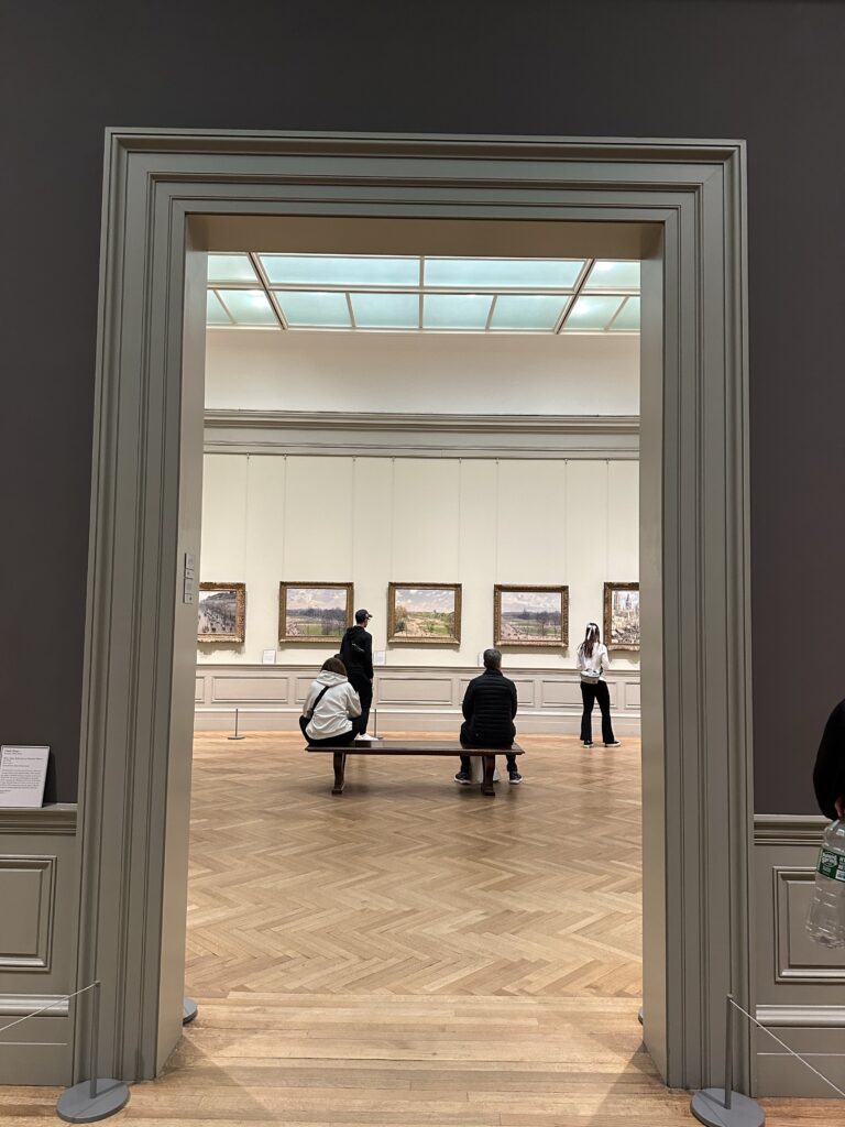 View through a wide doorway into a bright museum gallery with a skylit ceiling and herringbone wood floors. Four visitors explore the space, two seated on a central bench and two standing, while a row of gilt-framed Impressionist landscape paintings lines the white wall opposite.
Photo credit: Flora Whitehead via Dupe Photos