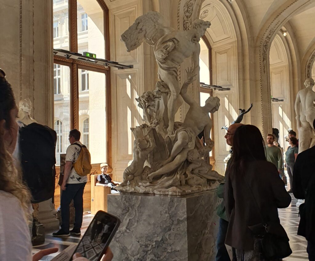 Visitors gather around a large dramatic white marble sculpture of intertwined figures on a gray marble pedestal, displayed in a sunlit grand gallery hall with ornate vaulted ceilings, arched windows, and marble tile floors. A visitor in the foreground photographs the work on a smartphone.
Photo credit: Gabs C via Dupe Photos