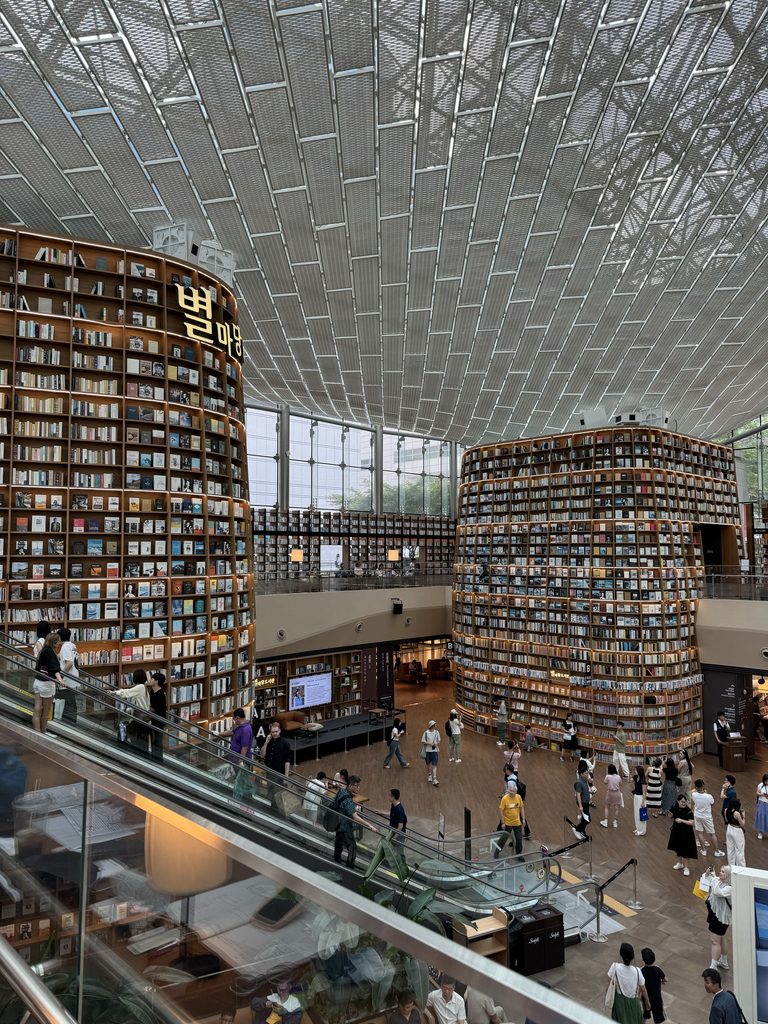 A vast modern library interior with towering cylindrical bookshelves filled with thousands of organized volumes, under a sweeping latticed ceiling. Visitors browse and move through multiple levels connected by escalators, illustrating the challenge and opportunity of making large, well-organized collections accessible and navigable to the public. Photo credit: Barbara Grigoryeva via Dupe Photos