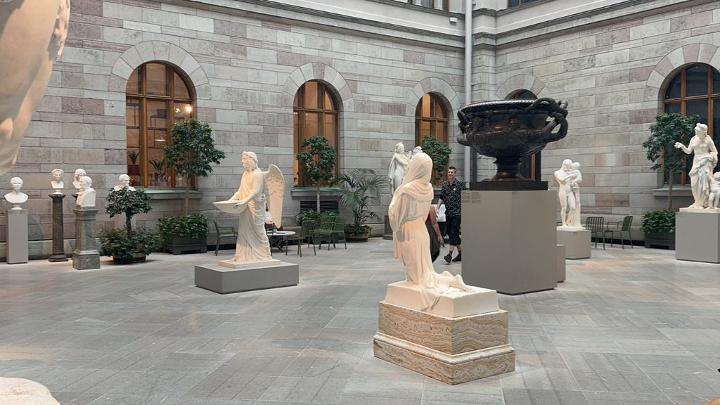 A museum sculpture courtyard displaying multiple white marble figural works and a large bronze urn on individual pedestals, surrounded by arched stone walls and potted trees. The open, navigable layout reflects how well-connected physical collections invite exploration, the same principle that should guide how museums structure their collections, content, and audiences online. Photo credit: Sophie via Dupe Photos