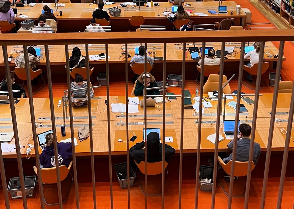 Researchers and students work across multiple levels of a modern library reading room, surrounded by floor-to-ceiling bookshelves filled with organized collections. The scene reflects the kind of structured, purposeful knowledge management that effective museum digital transformation should support, where technology serves the work rather than complicating it. Photo credit: Daniela Lara via Dupe Photos