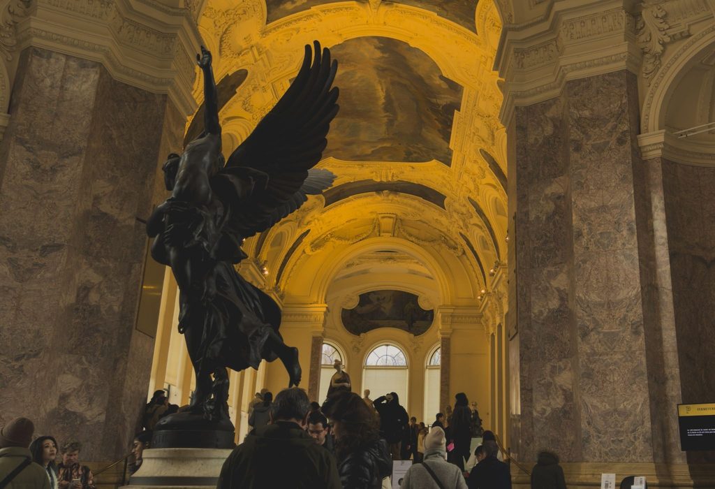 Visitors gather beneath a large dark bronze winged sculpture in the grand vaulted entrance hall of a historic museum, with ornate Baroque arches, ceiling murals, and stained glass windows stretching into the distance. The scale and grandeur of the physical space reflects the rich institutional knowledge museums are now being asked to bring into the digital world. Photo credit: Chloe G via Dupe Photos
